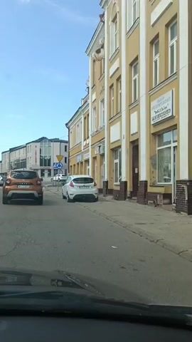 This video captures a view from inside a vehicle driving through a town. The scene is an urban street lined with buildings, featuring a clear blue sky and bright sunlight, suggesting it's daytime with good weather.

The primary subjects are the buildings and the vehicles on the street. The building on the right is a yellow, two-story structure with a white trim and several windows. Signs are visible on the building's facade, including one with blue lettering that reads "GEMDEZJA" and another indicating "USŁUGI PROJEKTOWE". To the left, a modern building with glass windows is visible in the distance.

Several cars are present: an orange SUV in the foreground, a white hatchback parked on the right side of the street, and other vehicles in the background. Two women are walking on the sidewalk in front of the yellow building, one wearing a patterned dress and the other a striped shirt and light-colored pants.

Road signs are also prominent. A blue circular sign with a white arrow indicating a roundabout is visible, as is a pedestrian crossing sign with a blue background and a white pedestrian symbol.

The camera perspective suggests the video is taken from the driver's seat or a passenger seat of a moving car, as the windshield and dashboard are partially visible at the bottom of the frame. The activity is simply driving through a town street. The location context indicates this is Ostrów Mazowiecka, Poland.