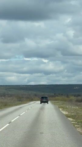 The video shows a dark sedan traveling on a gray asphalt road through a hilly landscape under a cloudy sky. The road stretches straight ahead with a solid white line separating the lanes. The surrounding terrain consists of low, green hills and scattered vegetation. The sky is predominantly overcast with dark, rolling clouds. The camera is positioned behind the moving car, capturing its rear view as it drives away.