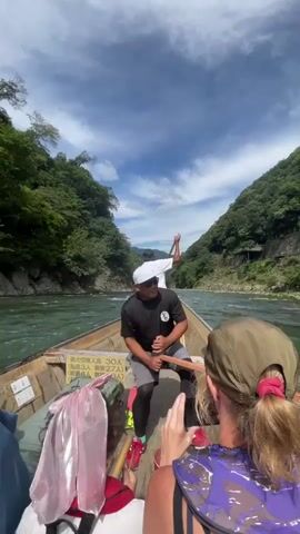 A perspective shot from a boat moving down a river. Lush green trees line the riverbanks, and the sky above is partly cloudy. In the foreground, three people are visible from behind, two women wearing hats and a child. A man wearing a white hat, sunglasses, a black t-shirt, and shorts is in the center of the boat, holding an oar and appearing to steer or guide the vessel. Another person is visible in the background, also holding an oar. The boat has wooden sides and appears to be carrying passengers. A sign with Japanese writing is attached to the side of the boat. The water is a greenish-blue, and the river is flowing. The overall scene is one of outdoor recreation and tourism.