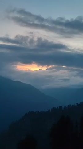 The video captures a mountain landscape at dusk or dawn. The sky is filled with dramatic, dark clouds, with a sliver of orange light peeking through, suggesting either sunrise or sunset behind the mountain peaks. Layers of mountains recede into the distance, with the foreground featuring a dark, silhouetted treeline. The overall mood is serene and atmospheric. There are no people or specific objects visible in the frame.