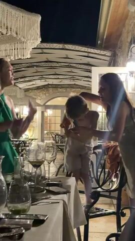 A young boy, shirtless and wearing light-colored shorts, is dancing with enthusiasm at an outdoor dining table. He is positioned between two women, one on his left wearing a green dress and the other on his right whose face is not fully visible. The woman in the green dress is also dancing and appears to be singing, with her arms raised. The table is set with white tablecloths, wine glasses, and what appear to be pitchers of water or beverages, suggesting a meal is taking place. The background shows an outdoor seating area with ornate metal chairs and arched architectural elements, illuminated by soft lighting. The scene takes place at night.