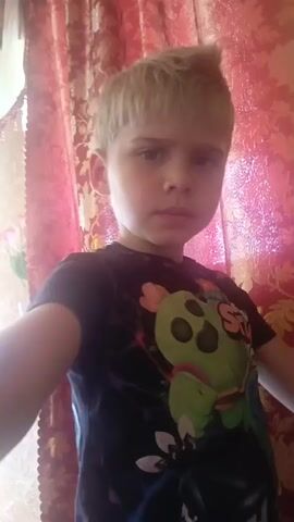 This media file is a short video clip featuring a young boy with blonde hair and blue eyes. He is wearing a black t-shirt with a graphic print that appears to be from the video game "Brawl Stars," depicting the character Spike. The background suggests an indoor setting, with pink curtains visible. The boy's expression is somewhat neutral, and he looks directly at the camera. The video is shot from a close-up perspective, focusing on the boy's face. Some segments of the video are out of focus or blurred, possibly due to camera movement or the filming process. There is no discernible activity or event taking place beyond the boy posing for the camera. The time of day and weather conditions are not evident from the visual information. The location context provided is Oryol, Russia.