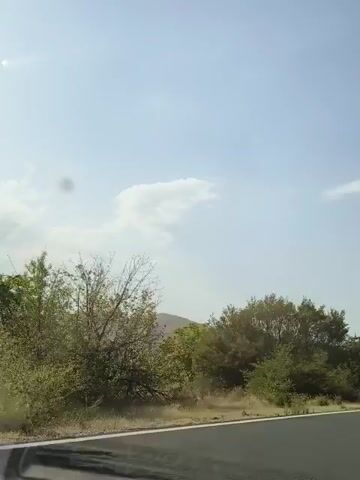 The video shows a view from a moving vehicle on a paved road. The sky is bright blue with a few scattered clouds. To the left of the road are trees and vegetation, with some barren hills in the distance. To the right of the road, there are more trees and what appear to be solar-powered streetlights. The road itself has white lane markings. The camera is angled slightly downwards towards the road and upwards towards the sky, giving a sense of movement and forward momentum.