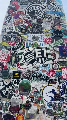 The video shows a surfboard covered in stickers, standing upright on a beach. The surfboard is mostly white with various colorful stickers of different shapes and sizes. Some of the stickers are branded with logos and text, while others are illustrations or photos. In the background, there is the ocean and a cloudy sky. The camera pans up and down the surfboard, showcasing the multitude of stickers. No people are visible in the video, and no actions or interactions are occurring. The environment is a typical beach scene with the ocean and pebbles on the ground.