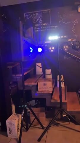The video shows a dimly lit indoor space with several light fixtures set up on stands. The lights are pulsing and changing colors, primarily blues and purples, with some white and yellow lights visible. In the background, there are wooden steps with various objects on them, including what appear to be white buckets and a wooden crate. A blackboard with writing and drawings is visible on the wall behind the lights. A television screen is also present. The overall atmosphere suggests a setup for a party or event.