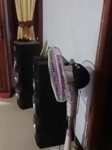 The video opens with a static shot of a room interior. To the left, a dark wooden door with a silver handle is visible. Next to it, a large, tall, black speaker stands against a light-colored wall. To the right of the speaker, another similar black speaker is positioned. In front of these speakers is a white and purple oscillating fan. The wall behind the speakers has a yellow curtain draped over a window.

The video then transitions to a shot where the wooden door is slightly ajar, revealing a glimpse of the outside. A red object, possibly a shoe, lies on the floor near the door. The view shifts to show a motorcycle parked outside.

The scene continues to pan, showing the open doorway more clearly. The motorcycle is now more visible, parked on a concrete surface. A portion of the house's exterior is visible, including a wooden structure and some foliage in the background. The video then shows another motorcycle parked closer to the camera on the left side, with its rear wheel visible. The wooden door is now fully open, providing a wider view of the outdoor area. The scene then widens further, showing more of the outdoor space and the two motorcycles.