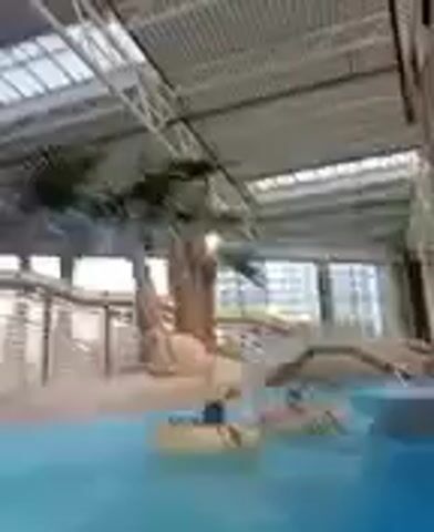 The video shows an indoor swimming pool complex. The pool is filled with bright blue water, and there are people floating in inflatable tubes. In the background, there is a decorative palm tree and a bridge. The ceiling of the complex is made of glass and metal, allowing natural light to filter in. The overall impression is one of a fun and relaxing environment.