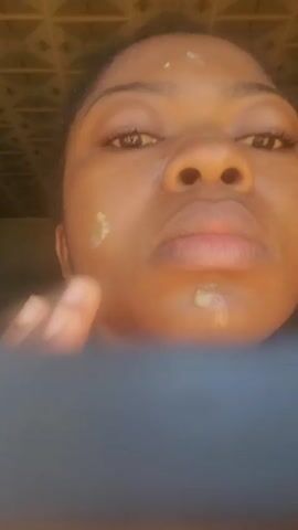 A person with dark skin and dark hair looks directly at the camera. White dots are visible on their forehead and a small amount of white substance is on their lower lip. They bring their hands up to their face and rub their forehead and temples, then move their hands down towards their cheeks. Their eyes are open, and their mouth is slightly open, revealing a gap between their front teeth. The lower portion of the frame is obscured by a dark, blurry object. The background is a blurry light-colored, possibly textured surface.