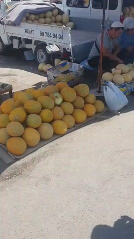 A street market scene unfolds with a focus on a display of melons. In the foreground, a young boy stands facing the camera, wearing a patterned t-shirt and striped shorts. To his left, a large pile of yellow melons is arranged on a mat. Behind the boy, a man in a grey shirt and a woman in a blue headscarf are seated, appearing to be vendors, with more melons stacked around them. A white truck is parked in the background, also laden with melons. A woman in a tie-dye shirt is visible on the right side of the frame, facing the seated vendors. The ground is paved, and the overall atmosphere suggests a busy outdoor market.