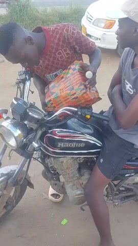 This video, recorded in Picha Ya Ndege, Tanzania, shows two young men gathered around a motorcycle. One man, wearing a red t-shirt with a repeated monogram pattern, is leaning over the motorcycle, holding a colorful, woven bag. He appears to be pouring something from the bag into the motorcycle's fuel tank. The other man, wearing a grey tank top and a beige baseball cap, sits on the motorcycle, observing the activity with his arms crossed.

The setting appears to be outdoors on a dirt road, with a white car and a truck with a blue tarp visible in the background, suggesting a roadside or parking area. The sky is visible with some clouds, indicating it is daytime. The overall mood seems casual and perhaps a bit practical, as they are tending to the motorcycle.

The motorcycle itself is a prominent object, a dark-colored model with a visible engine. Text is visible on the motorcycle's fuel tank: "HAOJUE". A green object, possibly a piece of trash, lies on the ground near the motorcycle's kickstand. The presence of the vehicles and the outdoor setting suggest a possibly rural or semi-urban environment in Tanzania.