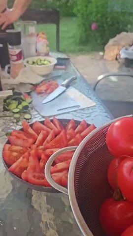 The video shows a table set for an outdoor meal, likely a salad preparation. There are several bowls and plates filled with chopped cucumbers and sliced tomatoes. A bottle of olive oil, a salt grinder, and a knife are also visible on the table, suggesting ingredients for a salad dressing. In the background, a person's hands are visible, possibly engaged in food preparation. The setting appears to be a backyard or garden, with greenery and other outdoor elements in the background. The lighting suggests it is daytime.