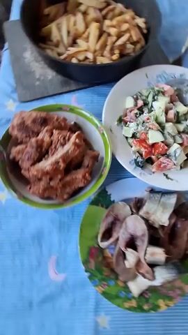 A table is covered with a blue and white striped tablecloth. On the table are several dishes of food. In the upper left corner is a black frying pan filled with french fries. To the right of the frying pan is a white plate with a salad of chopped tomatoes, cucumbers, and greens, mixed with a creamy dressing. Below the frying pan and to the left is a green and white bowl filled with pieces of fried meat. Below the salad plate is a smaller green plate with slices of fish. The lighting suggests it is daytime and the setting appears to be indoors. The camera is moving slightly, giving a sense of looking at the food from a close-up perspective.