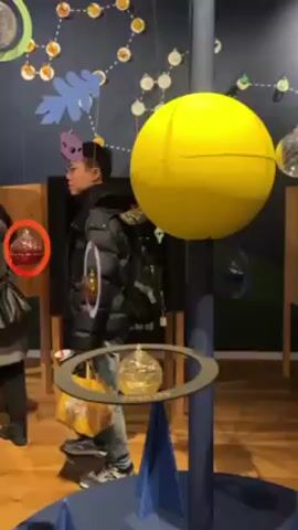 The video shows a person looking at an educational display about the solar system. The display features a large yellow sphere representing the sun. Hanging from the ceiling are models of the planets. Labels such as "Venus" appear to identify the planets. The room has a dark blue wall with constellations illustrated on it. A person in a black jacket and jeans is standing next to the display.