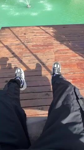 The video begins with a first-person perspective, showing the viewer's legs clad in black cargo pants and white and black sneakers. They are sitting on a wooden deck overlooking a vibrant green body of water. In the distance, a fountain sprays water upwards. As the video progresses, the view pans out, revealing a picturesque scene.

The setting is a resort or a similar establishment, characterized by several thatched-roof cabanas situated around the tranquil water. These cabanas have unique, circular openings and appear to be overwater bungalows or dining areas. The water is a striking emerald green, and the fountain in the center adds a dynamic element.

The time of day appears to be late morning or early afternoon, indicated by the bright sunlight casting distinct shadows of the viewer and some surrounding foliage onto the wooden planks. The weather seems to be pleasant and sunny, with a clear blue sky visible above.

The overall mood is relaxed and serene, suggesting a vacation or leisure activity. The presence of lush green bushes on the left side of the frame adds to the natural beauty of the location. The camera subtly captures the environment without focusing on any specific individuals, emphasizing the experience of being in this tranquil place.

No visible text is present in the video. The location context of Tambon Pa Kae Bo Hin, Thailand, further enhances the understanding of this tropical, resort-like setting. The video conveys a sense of peaceful enjoyment and appreciation for the scenic surroundings.