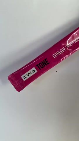 A pink stick pack labeled "TONE" and "БОЛЬШЕ ЭНЕРГИИ!" (More Energy!) is held by a person with red-painted fingernails against a white background. The stick pack appears to contain a powdered drink mix, and the packaging has a glossy finish. The person's hand is out of focus in the upper right corner of the frame. The background is smooth and white, suggesting a clean surface or tabletop.