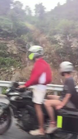 A daytime video captures two individuals riding a motorcycle with saddlebags on a winding paved road through a lush, green, mountainous landscape in Tân Quang Commune, Vietnam. A small waterfall cascades down a rocky hillside directly beside the road, which is bordered by a white guardrail. The sky is overcast, and the video is filmed from the perspective of a following vehicle, showing the scenic journey.