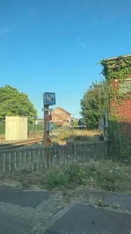 The video shows a blue sky and a train station. In the foreground, there is a fence and overgrown grass. A wooden pole with an SOS phone and a blue sign with a white phone symbol is visible. Below the SOS sign, there is a red circular sign with a diagonal line and some text, and a grey box that appears to be the phone. In the background, there is a brick building with a red roof and windows, and beyond that, trees and a train approaching on the tracks. The train is blue and white. The overall environment appears to be rural or semi-rural.