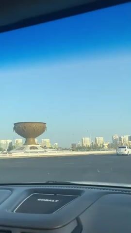 The video shows a view from inside a car driving on a road. The windshield of the car is visible in the foreground. In the distance, there is a city skyline with several tall buildings. There is also a large, ornate golden bowl-shaped structure on the left side of the road. The sky is clear and blue.