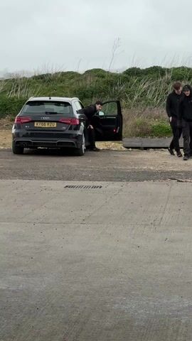 The scene shows three individuals near a black Audi A3 parked on a paved area. One person is exiting the driver's side of the vehicle, while the other two are walking away from the car towards the right side of the frame. The car has a license plate reading KT66 RZU. The surrounding environment consists of a concrete or paved surface, some grassy vegetation, and an overcast sky. The setting appears somewhat rural or industrial.