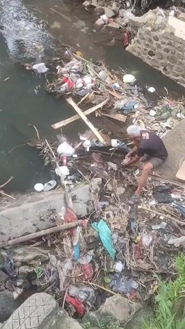 A medium shot shows a man in a black shirt and dark shorts squatting near a trash-filled canal. He has gray hair and is crouched in front of a pile of debris, seemingly sifting through it with his hands. The canal is filled with a variety of trash items, including plastic bags, bottles, containers, and organic debris like branches and leaves. Some trash is also piled on the bank of the canal. The canal walls are made of concrete and stone. There is some green foliage visible on the far side of the canal.