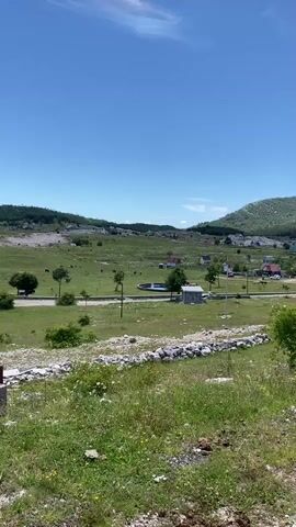 The video shows a wide shot of a rural landscape on a clear sunny day. In the foreground, there is a patch of green grass with some stones and dirt. A low stone wall runs horizontally across the middle ground, separating the foreground from a grassy field. Beyond the wall, there is a gently rolling green landscape dotted with scattered trees and houses, many of which have red roofs. In the mid-ground, there is a circular blue pond or pool. A small, white, shed-like structure is visible near the pond. Further in the distance, a large, tree-covered hill forms the backdrop, with more houses nestled on its slopes. The sky is a clear, bright blue with a few wispy white clouds. The overall impression is one of a peaceful, undeveloped countryside. There are no people or animals clearly visible in the video.