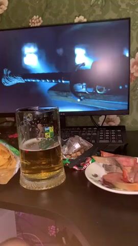 A close-up shot shows a table with a glass of beer, a bag of potato chips, and a plate of snacks in front of a computer monitor. The monitor displays a scene from what appears to be a video game or movie, with bright blue lights and a dark, futuristic vehicle. The beer is in a large glass mug with a logo. The bag of chips is open, and some chips are visible. The plate of snacks contains what looks like dried meat and nuts. The background is a wall with green floral wallpaper. A keyboard is visible behind the monitor. The lighting is dim, with some colorful light coming from the bottom left of the frame, possibly from a computer tower.