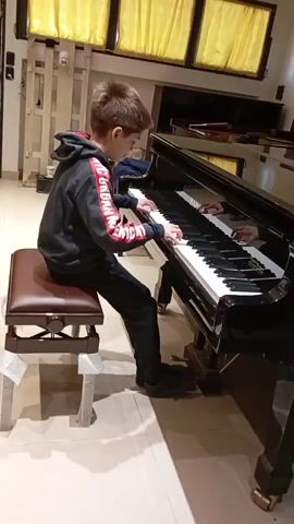 A young boy is sitting on a piano bench playing a black grand piano. He is wearing a black hoodie with red and white writing on the sleeves and black pants. The piano is in a room with tiled flooring. Yellow curtains hang in the background.
