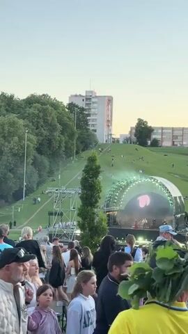 The media file shows an outdoor concert or festival scene in Jonava, Lithuania. A large crowd of people is gathered, looking towards a stage with a prominent arched structure and lighting. Performers are visible on the stage, and the atmosphere appears festive. In the background, a lush green hill is dotted with people relaxing and watching the event. Several buildings are visible on top of the hill and further in the distance, suggesting an urban or semi-urban setting. Tall trees are interspersed throughout the scene, particularly around the stage area and along the side of the hill.

The time of day appears to be late afternoon or early evening, with soft, warm lighting and a clear sky. The weather seems pleasant for an outdoor gathering. The overall mood conveyed is one of enjoyment and community, with people engaged in watching the performance and socializing. The presence of a large audience and a professionally set up stage indicates a significant public event. No visible text is present in the media file.