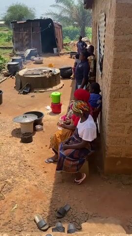 The video depicts an outdoor scene near a brick building. Several individuals are present. In the foreground, three women are seated on a low wall or bench, facing away from the camera. They are dressed in traditional attire, with head coverings. One woman in the foreground wears a white shirt and a colorful patterned skirt. To their right, a group of people, including men and women, are standing.

In the middle ground, several large cooking pots are placed around a well-like structure. Fires appear to be lit under some of the pots, suggesting cooking is in progress. A woman in a blue shirt and black pants is actively engaged with one of the pots, stirring its contents. Another woman, in the background, is also tending to the cooking area. Two green and red containers are stacked near the well. Various buckets and containers are scattered around the cooking area.

The environment appears to be somewhat rural or semi-urban, with dirt ground, some vegetation in the background, and a corrugated metal structure to the left. The building to the right is made of brick and has a window covered with corrugated metal. Several pairs of sandals are visible on the ground in the foreground. The lighting suggests it is daytime. The overall impression is of a community preparing or cooking food outdoors.