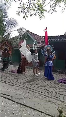 The video shows a line of people, likely children, dressed in elaborate costumes and headdresses. They appear to be participating in a parade or performance. The costumes feature large, decorative elements such as feathers, wings, and ornate headpieces. The people are standing on a paved surface, possibly a street or walkway. In the background, there is a house with a red and white flag flying. Trees and foliage are visible, suggesting an outdoor setting. The overall impression is one of a cultural event or celebration.