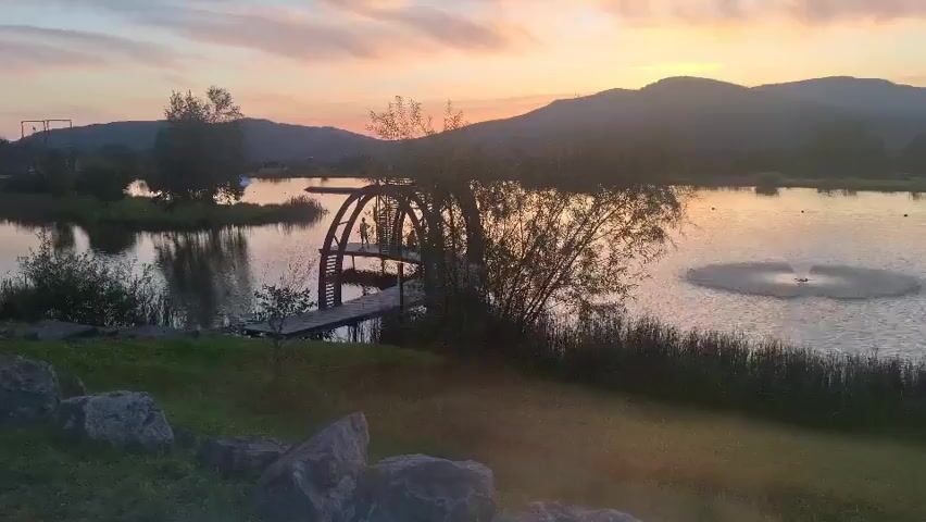 A serene sunset over a tranquil lake. The sky is painted with soft hues of orange and pink, with streaks of clouds drifting across. In the foreground, lush green grass slopes down towards the water, dotted with large grey rocks. A wooden walkway leads to a unique arched structure on the lake, resembling a small dock or platform with an arched trellis. Reeds and bamboo plants line the edge of the water, adding to the natural beauty of the scene. In the distance, rolling hills are silhouetted against the twilight sky. A circular fountain in the lake creates a gentle spray, adding a touch of movement to the peaceful landscape. There are no people or animals visible in the video.