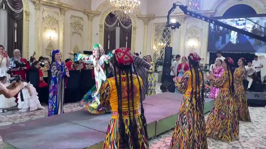 This is a video clip of a cultural performance or celebration, likely taking place in Bekabad, Uzbekistan. The scene is set in an opulent, indoor venue, suggested by the gilded ornate walls, large crystal chandelier, and luxurious draperies. The floor is carpeted, and a raised platform, possibly a stage or runway, is set up in the foreground.

The central activity features a group of individuals, predominantly women, dressed in vibrant, traditional clothing. The attire is characterized by colorful, intricately patterned fabrics, long flowing sleeves, and distinctive headwear, including embroidered caps and headscarves. Several women are dancing or posing on the platform, their movements suggesting a choreographed performance. Their expressions appear joyful and engaged, conveying a festive atmosphere.

Among the performers, one woman in a particularly striking, multi-colored embroidered dress and a green headscarf is a focal point. Another woman in the foreground wears a bright yellow top with a red, intricately patterned headpiece and long braids. A man in traditional attire, possibly a dance leader or participant, is also visible on the platform.

The audience is present, with some individuals in the background holding up phones, likely recording the event. One person in the foreground, wearing a blue vest with text "BOOM AUDIO STUDIO", appears to be operating a camera. The lighting is bright, suggesting it is daytime or a well-lit indoor space. The overall mood is celebratory and rich with cultural expression.