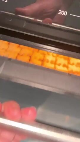The video shows a close-up view of a baking rack filled with numerous identically shaped cookies, possibly made in a bear cookie cutter. The cookies are golden brown, indicating they are freshly baked and are arranged in neat rows on what appears to be parchment paper. They are inside an oven, with the oven light illuminating them. The oven interior, including the rack below and the door's glass, is visible, reflecting the light and the cookies. The shot is taken from a slightly elevated angle, looking down into the oven. There is no indication of time of day, weather, or specific location cues beyond what can be inferred from the oven itself. The overall impression is of a home baking activity. There is no visible text in the video.
