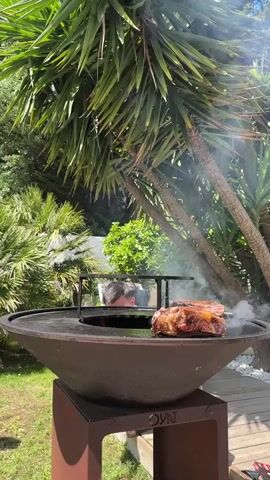 The video shows a man grilling meat on an outdoor grill. The grill is a round, bowl-shaped structure with a flat cooking surface and a fire pit in the center. The man is wearing a white T-shirt and white shorts. He is standing near the grill, looking at the meat. The meat is a large cut of beef, possibly a ribeye steak. There is smoke rising from the grill. The background includes tropical trees and vegetation. The grill is labeled "OVNI."