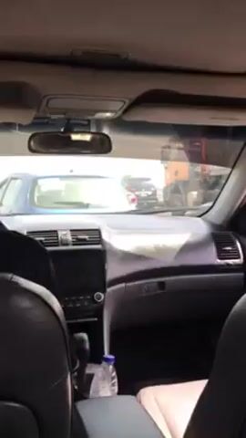 A car's interior is viewed from the back seats, looking forward through the windshield. A driver is visible in the front seat, wearing a dark top. The dashboard is gray, and a small screen is visible in the center console. A water bottle sits in a cup holder. Outside the car, a line of other vehicles is visible, including cars and a large orange truck. The scene appears to be a traffic jam. The speaker says "enter", "enter", "enter".