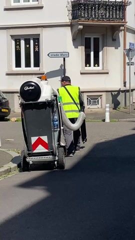 The video shows a man wearing a yellow safety vest and a black hat walking and pushing a motorized trash can through a street. The trash can is black and white and has the word "Glutton" written on it. A bottle of water is visible attached to the side of the trash can.  A sign labeled "stade Mars" is visible behind the man. A black car and white bollards are visible to the left of the man. A white building with windows is visible in the background. The street is paved. The video is recorded in broad daylight.