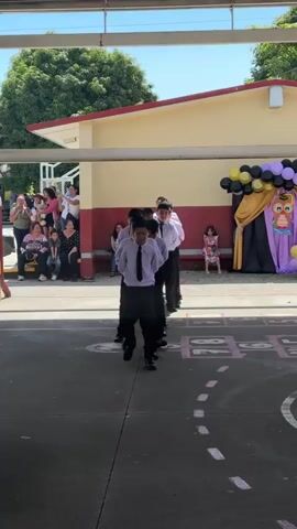 A group of young boys in white shirts and black ties and pants are standing in a line on a paved surface, possibly outdoors under a covered walkway. They appear to be performing a synchronized routine. In the background, there are spectators sitting and standing, a building with yellow and red siding, trees, and a decorated backdrop with balloons and an owl cutout. A young girl in a pink dress is sitting on the ground near the backdrop. The overall impression is of a school event or performance.