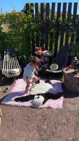 A young child is playing outside on a sunny day. The child is standing on a large, plush panda rug that is placed on a pink fluffy rug. The child is wearing a light blue t-shirt and white shorts. The child is reaching out towards a wooden stump with a black bird figurine on it. Behind the child, there is a wooden fence covered in green ivy, and two empty chairs. There are also some toys scattered on the ground and a small table with more toys on it. The ground is covered with gravel.