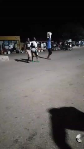 A nighttime scene unfolds in what appears to be a rural or semi-urban area. The lighting suggests it might be after sunset, with artificial lights illuminating the surroundings. Three individuals are the central focus. The person on the far left is partially obscured, walking towards the right. Next to them, a second person, also walking to the right, carries a green object, possibly a broom or a mop, which is bent. The third person, positioned further to the right and slightly ahead of the other two, is carrying a white container, possibly a bucket, balanced on their head. This individual is also walking to the right. In the background, there are several structures that resemble market stalls or small shops, with some activity or people visible near them. Several motorcycles are parked or moving in the background, indicating a mode of transportation common in the area. The ground is paved, appearing to be a road or a large open area. The lighting casts distinct shadows on the ground, some of which are quite long, suggesting the light source is low and to the left. A fourth person, a woman wearing a white head covering, a white t-shirt, and black leggings, walks towards the viewer from the right side of the frame, her body angled slightly away. She is looking down and appears to be holding something near her mouth, possibly a mask or touching her face. She is wearing flat sandals. To the far right of the frame, a portion of a three-wheeled vehicle, likely a tuk-tuk, is visible. The overall impression is one of everyday life continuing into the evening in a community.