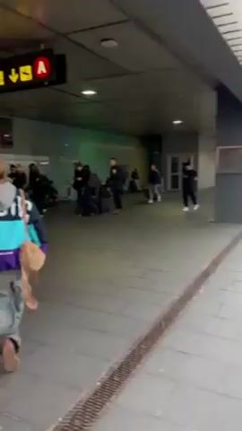 The video shows a bustling indoor public space, likely a transit station or airport terminal, with numerous people moving and stationary. The camera follows a person from behind, wearing a teal and purple jacket and carrying a tan-colored bag. People are seen carrying luggage, sitting on the floor, and walking in various directions. Overhead signs indicate directions, one marked "A". The flooring is tiled. The lighting is a mix of natural and artificial, creating a somewhat shadowed atmosphere. The scene conveys a sense of activity and transit.