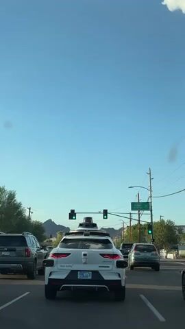 The video shows a white autonomous vehicle driving in traffic. The car is equipped with various sensors on its roof, including cameras and lidar. The traffic light is green, indicating that the car can proceed. Other vehicles are present in the scene, including SUVs and sedans, as well as a mountain range in the distance under a clear blue sky. The autonomous vehicle appears to be operating normally in a real-world driving environment.