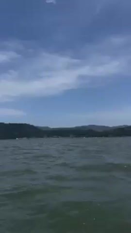 The video shows a view of the water from a boat. The water is choppy. In the background is a shoreline with trees and hills. The sky is blue with some white clouds. There is some ambient noise, possibly wind or the sound of the boat.