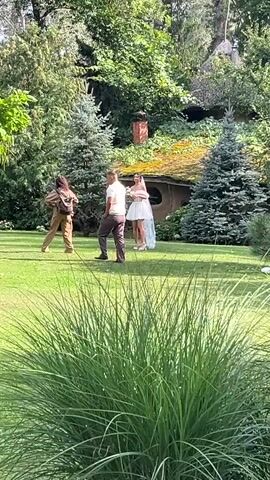 A video shows a wedding photoshoot in a garden. A bride in a short white dress stands in the center of the frame, holding a bouquet. To her left, a woman with a camera is taking her picture. To the bride's left and further back, a man in a light-colored shirt and dark pants walks away from the camera. In the background, there is a small, hobbit-like house with a moss-covered roof and a brick chimney. Lush green trees and bushes surround the scene.