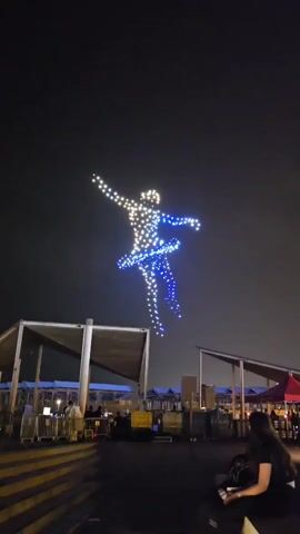 This video shows a drone light show at night. Hundreds of synchronized drones form the silhouette of a dancing ballerina in the sky. The ballerina is illuminated with white and blue lights. Below the drone display, there is a gathering of people watching the show. In the foreground, a woman with long dark hair is sitting and holding a phone, possibly recording the event. There are also structures that appear to be canopies or shelters in the background, and some lights indicating a public event or venue.