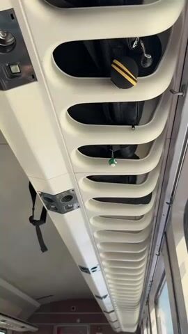 The video shows a close-up view of a luggage rack inside a train carriage. The rack is made of white plastic with a series of oval-shaped openings. Several black bags are stored in the rack. Two keychains hang from the bags. One keychain has a yellow and black striped item, and the other has a green and white billiard ball with the number 14 on it. The camera moves slightly to focus on the billiard ball keychain. The environment appears to be dimly lit, and the overall impression is one of a travel setting.