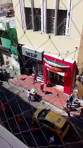 From an elevated perspective, the video captures a bustling street scene. The street is populated with numerous people, most of whom are pedestrians moving about. A yellow taxi cab is visible, and a person is pushing a cart down the street. Several shops line the street, including one named "Piko Riko," "La Conaza," and "Calzado Sk." Many vendors appear to have set up stalls or stands, indicated by the presence of umbrellas and other temporary structures. Overall, the scene suggests a vibrant and lively commercial district.