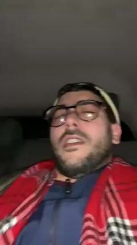 Here's a description of the video:

A man with glasses, a beard, and a white headband sits in the back seat of a car. He's wearing a blue jacket and has a red plaid blanket draped over him. He is singing along to music, but his eyes are mostly closed. The interior of the car is dark.