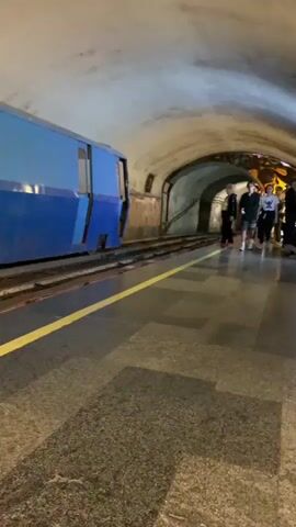 The video shows a blue subway train with doors open, but marked with a red and white tape, indicating it is out of service. The train is stationary in the station. A yellow line is painted on the platform to mark a safety boundary for passengers. The station has a curved, tunnel-like design with an arched ceiling. In the distance, two people stand on the platform. The overall atmosphere suggests an underground environment.