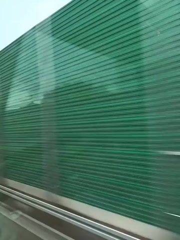 The video shows a view from a moving vehicle, possibly a train or car, looking out a window.  The foreground features a metallic rail or structure.  Immediately beyond the window is a tall, green, corrugated barrier, likely a sound barrier, which extends across the entire frame.  Beyond the barrier, the environment is visible.  In the first few seconds, the landscape is mostly obscured by the barrier.  As the vehicle moves, glimpses of trees, green grass, and a blue sky with some clouds become visible.  In the latter part of the video, a white building with a tall cylindrical structure next to it can be seen, along with more trees and foliage. The overall impression is of a journey through a semi-urban or suburban area, with infrastructure like sound barriers alongside natural elements. The motion blur indicates the continuous movement of the vehicle.