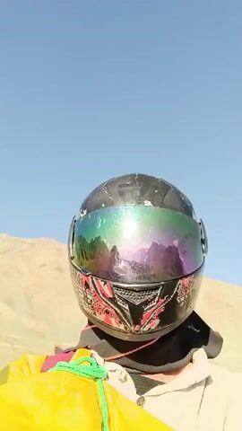 The video begins with a close-up shot of a person wearing a black motorcycle helmet with a rainbow-tinted visor and a colorful decal on the chin bar. The person is wearing a light-colored jacket over a darker garment, and a yellow bag with a green rope is visible in the foreground. The background is a barren, sandy landscape with mountains under a clear blue sky. As the video progresses, the camera perspective shifts, showing the person is on a motorcycle with another person also wearing a helmet riding behind. The motorcycle is traveling on a paved road, with more mountainous terrain and sparse vegetation on either side. The sky remains blue with a few wispy clouds. The footage appears to be filmed during daylight.